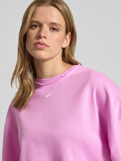 Frauenmodel trägt Cropped Sweatshirt mit Logo-Stitching von Nike in Pink, Relaxed-Fit, Rundhalsausschnitt, elastische Ärmelabschlüsse, Nahaufnahme Oberkörper.