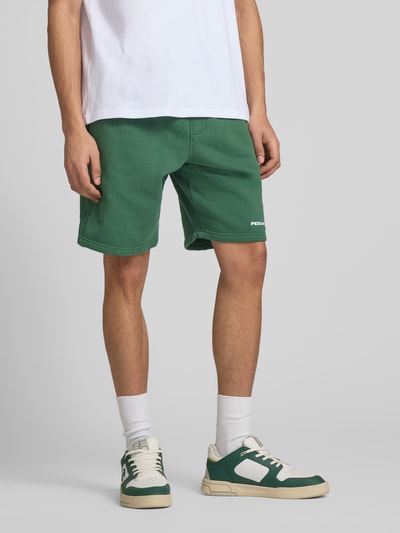 Męskimodel nosi szorty mit elastischem Bund und Logo-Stitching von Pegador, zielony, mieszanka bawełny, krój loose fit, wpuszczane kieszenie, szerokość w rozmiarze M 38 cm, długość wewnętrzna nogawki 23 cm, Ganzkörperansicht von vorne.