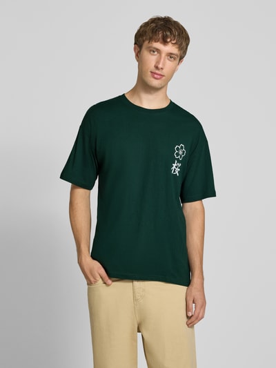 Männermodel trägt Relaxed Fit T-Shirt aus reiner Baumwolle Modell "DYLAN" von JACK & JONES in Dunkelgrün mit Print-Detail, Nahaufnahme Oberkörper. Rückenlänge bei Größe M beträgt 70 cm.