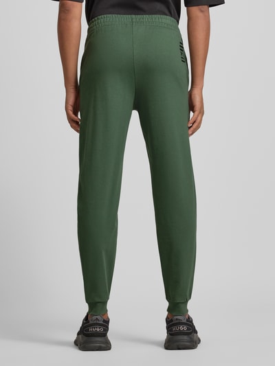 Männermodel trägt grüne Sweatpants mit Logo und elastischem Bund von EA7 Armani aus Baumwoll-Elasthan-Mix, Regular Fit, Ganzkörperansicht Rückansicht.