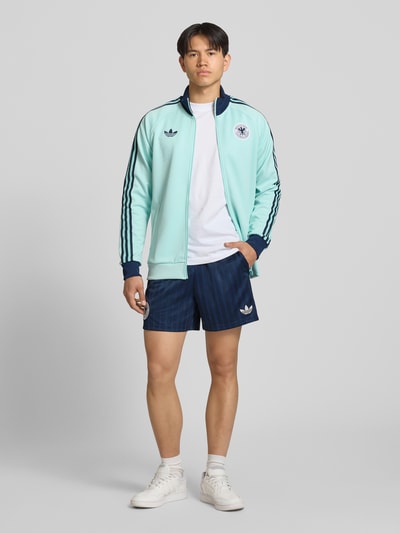 Męski model nosi Loose Fit Trikot-Shorts mit DFB-Applikation von adidas Originals, granatowy jacquard z wzorem, elastyczny pas, krój loose fit, boczne kieszenie, logo, kontrastowe paski, widok całej sylwetki z przodu.