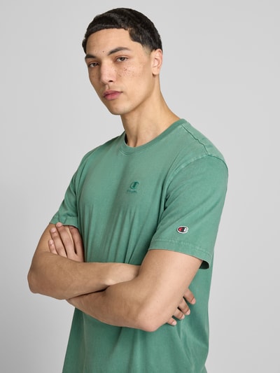 Męski model nosi T-Shirt mit Label-Stitching und Rundhalsausschnitt od CHAMPION z bawełny, krój regular fit, długość tyłu w rozmiarze M: 67 cm, zielony kolor, prosty fason, gładka faktura. Nahaufnahme Oberkörper.