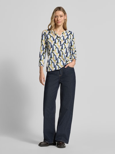 Vrouwenmodel draagt t-shirt met 3/4-mouw model 'FELICITY' van Soyaconcept, katoen- en viscosemix, regular fit, v-hals, ruglengte maat S 63 cm, geometrisch patroon in blauw, geel en wit, vooraanzicht volledige lichaamsopname.
