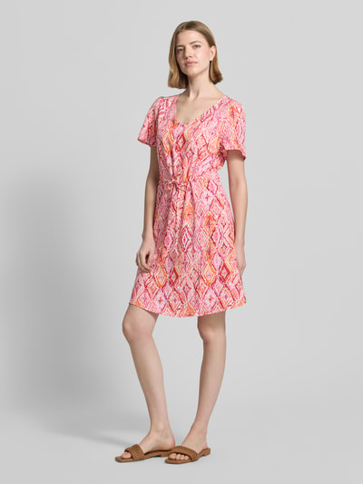 Frauenmodel trägt tailliertes Freizeitkleid aus reiner Viskose Modell 'EASY JOY' von Vero Moda mit Allover-Muster in Rosa und Orange, Mini-Länge, V-Ausschnitt, Kurzarm, glatte Textur, Ganzkörperansicht von vorne.