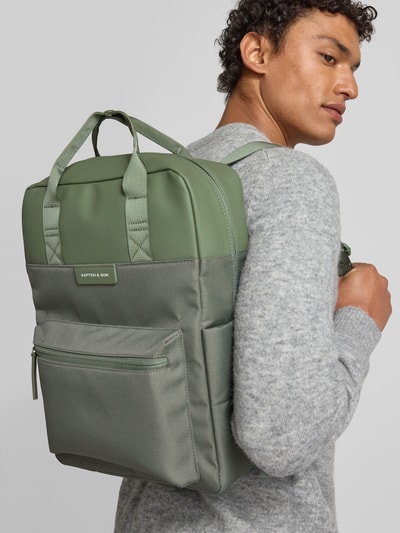 Männermodel trägt Rucksack mit Label-Detail von Kapten & Son in grünem Material-Mix, Hauptfach mit Reißverschluss, Schulterriemen, Nebenfach, Ganzkörperansicht Rückansicht.