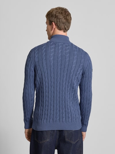Männermodel trägt Strickpullover aus reiner Baumwolle von Superdry in Blau mit Zopfmuster, gerippte Abschlüsse, Rückenlänge 70 cm, Ganzkörperansicht Rückansicht.