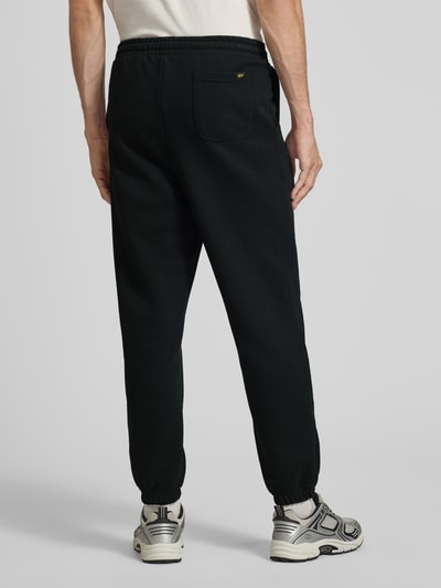 Männermodel trägt Sweatpants mit Label-Print von Alpha Industries, schwarze Farbe, lockere Passform, Baumwoll-Mix, Ganzkörperansicht Rückansicht.