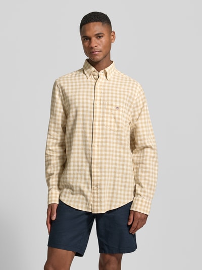 Męski model nosi Regular Fit Leinenhemd mit Baumwoll-Anteil Modell 'GINGHAM' od Gant z mieszanki lnu i bawełny, żółto-beżowa krata, kołnierzyk typu button down, wyhaftowane logo na brustaszy, długa listwa guzikowa, krój regular fit, długie rękawy, zaokrąglony dół, długość tyłu w rozmiarze M: 66 cm, szerokość w rozmiarze M: 46 cm, długość rękawów w rozmiarze M: 64 cm, całkowity widok od przodu.