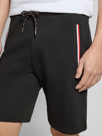 Mężczyzna nosi Shorts mit elastischem Bund von Christian Berg Men, czarny bawełniano-elastanowy krój regular fit, kieszenie z kontrastowym paskiem, elastyczny pas z tunelem, szerokość w rozmiarze M: 43 cm, długość wewnętrzna nogawki w rozmiarze M: 23 cm, zbliżenie na dolną połowę ciała
