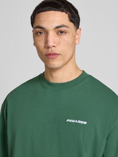 Męski model nosi Oversized T-Shirt mit Logo-Stitching von Pegador, zielony, bawełna, krój oversized fit, okrągły dekolt, wyhaftowane logo, długość tyłu w rozmiarze M: 73 cm, zbliżenie na górną część ciała.