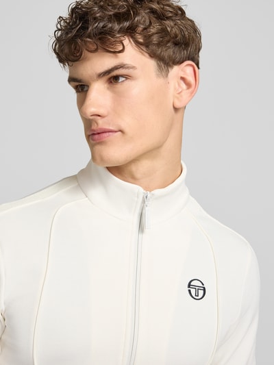 Männermodel trägt Slim Fit Tracksuit mit Logo-Stitching Modell 'Egeo' van SERGIO TACCHINI in gebroken wit katoenmix met gestikt logo, nahaufnahme bovenlichaam. Context: heren tracksuit, elastische katoenmix, slim fit, opstaande kraag, ritssluiting, elastische boorden, bandwijdte maat M: 42 cm, ruglengte: 64 cm, schouderbreedte: 46 cm, mouwlengte: 67 cm, binnenbeenlengte: 73 cm.