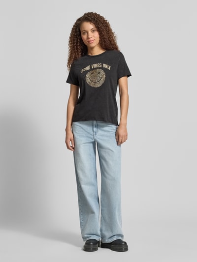 Frauenmodel trägt Regular Fit T-Shirt aus reiner Baumwolle Modell 'LISA LIFE LUCY' von Only in Schwarz mit Motiv-Print, unifarben, Rundhals, kurzarm, Jersey-Struktur, kombiniert mit weiter Jeans und schwarzen Schuhen, Ganzkörperansicht von vorne.
