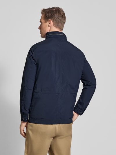 Męski model nosi Regular Fit Jacke mit Stehkragen od Bugatti w granatowym kolorze, aplikacja z logo, długość tyłu 72 cm, szerokość ramion 46 cm, szerokość w rozmiarze 52 cm, długość rękawów 68 cm, kieszenie na piersi, kaptur chowany, krój regular fit, widok całej sylwetki z tyłu.