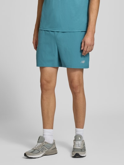 Männermodel trägt Shorts mit elastischem Bund und Logo von New Balance in Blau, Regular Fit, Nahaufnahme Unterkörper, Kontext: elastischer Bund, Tunnelzug, Logo, Bundweite M 39 cm, Innenbeinlänge 12 cm.