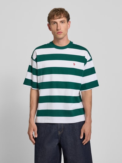 Männermodel trägt T-Shirt mit Label-Stitching und Rundhalsausschnitt von REVIEW, grüne und weiße Streifen, Reine Baumwolle, Relaxed-Fit, Allover-Look, Rückenlänge 67 cm bei Größe M, Ganzkörperansicht von vorne.