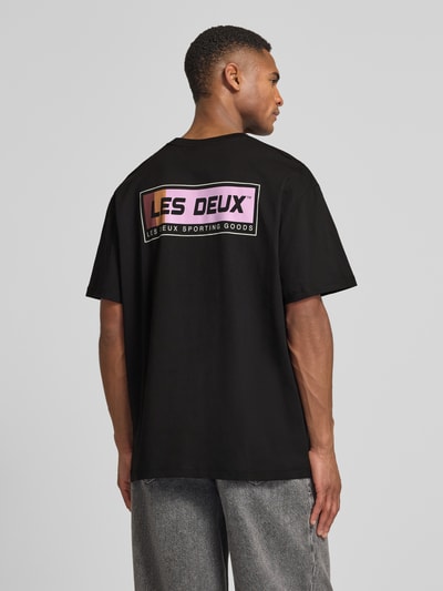 Mężczyzna model nosi T-Shirt mit Logo-Print Modell 'Chase' von Les Deux, czarna bawełna, prosty krój, widok całej sylwetki z tyłu.