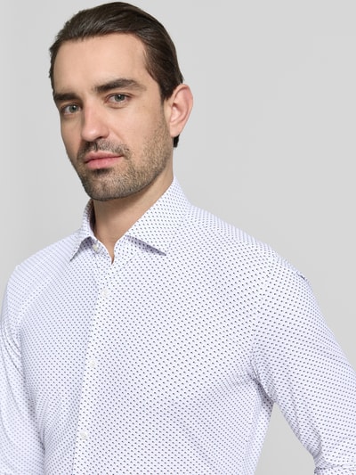 Mężczyznę model nosi Slim Fit Businesshemd mit Kentkragen Modell 'KENNO' von HUGO, efekt: wzór na całej powierzchni, kołnierzyk typu kent, wyhaftowane logo, pas na plecach, jednorzędowa listwa guzikowa, długi rękaw, zakończenie: zaokrąglony, mankiet, krój slim fit, kolor biały z ciemnoniebieskim wzorem, koszula biznesowa, zbliżenie na górną część ciała z przodu.