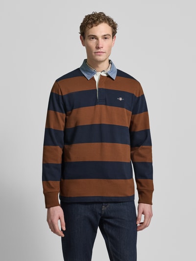 Męski model nosi Bluza z wyhaftowanym logo Sweatshirt mit Label-Stitching od Gant, bawełna, krój regular fit, kołnierzyk polo, listwa guzikowa, prążkowane zakończenia, długość tyłu 72 cm, długość rękawów 66 cm, paski brązowe i granatowe, gładka tekstura, widok całej sylwetki z przodu.