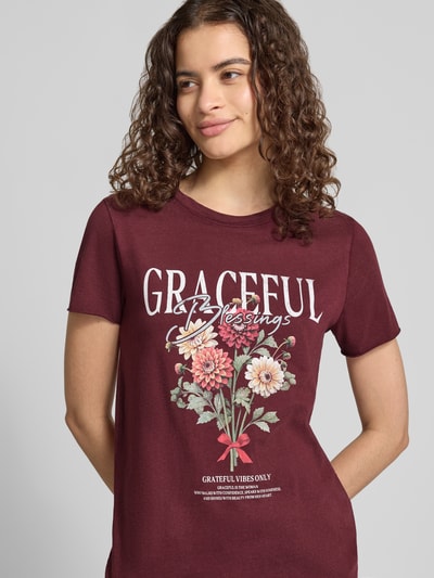 Frauenmodel trägt Regular Fit T-Shirt aus reiner Baumwolle 'ONLLUCY' von ONLY in Bordeaux mit floraler Grafik und weißem Schriftzug, Nahaufnahme Oberkörper. Damen T-Shirt von ONLY.