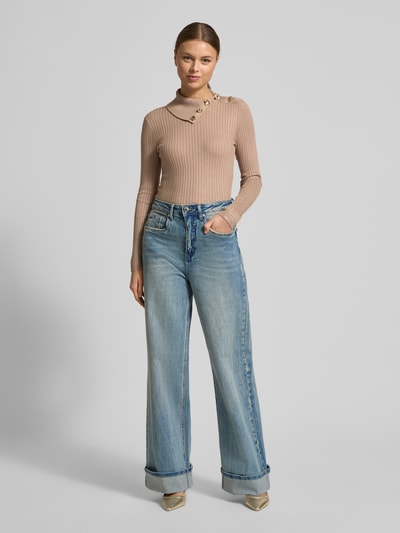 Modelka nosi Slim Fit Longsleeve aus Viskose-Mix Modell 'Sicilla' od Guess, beżowa dzianina, stójka, ozdobne detale, krój slim fit, widok całej sylwetki od przodu.