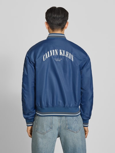 Männermodel trägt Regular Fit College-Jacke mit Label Stitching van Calvin Klein Jeans in look: effen met contrasterende details, lange mouw, college-kraag, ritssluiting, klepzakken, rompzoom en mouwboord geribd, applicatie met labelstitching, ganzkörperansicht rückansicht.