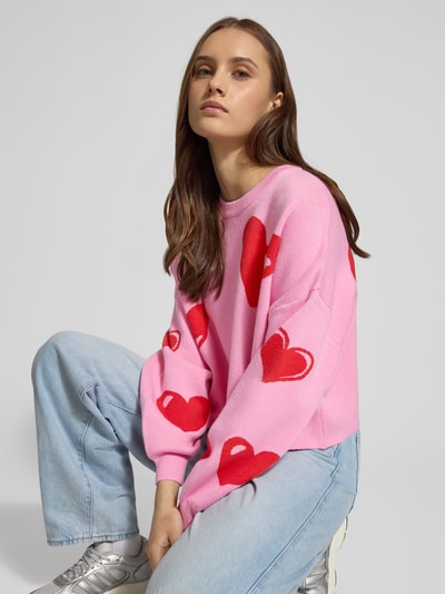 Frauenmodel trägt Oversized Strickpullover aus Viskose-Mix Modell 'SHYLA' von Noisy May, pink mit roten Herzen, Statement-Schriftzug, Ballonärmeln, O-Neck, Langarm, elastische Ärmelabschlüsse, Rückenlänge 44 cm, Ärmellänge 46 cm, Ganzkörperansicht von vorne.