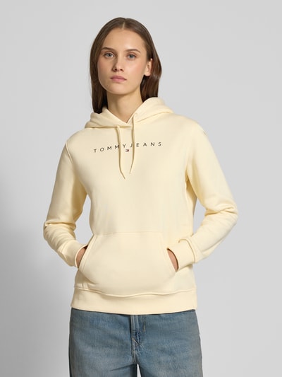 Modelka kobieca nosi Regular Fit Hoodie aus Baumwoll-Mix od Tommy Jeans w jednolitym beżowym kolorze, nadruk logo, długi rękaw, materiał sweat, widok na górną połowę ciała z przodu.