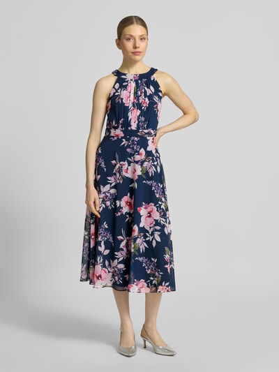 Frauenmodel trägt Midikleid mit floralem Allover-Print von Christian Berg Cocktail in Blau mit Pink-Tönen, Tailliert, Rundhalsausschnitt, Gelegte Falten, Ärmelloses-Design, Nahtreißverschluss, Ganzkörperansicht von vorne. Rückenlänge Größe 36: 115 cm, Brustweite Größe 36: 40 cm.