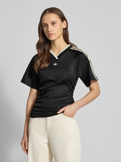 Vrouwenmodel draagt T-Shirt mit V-Ausschnitt van adidas Originals in zwart met asymmetrisch design, aansluitende fit, korte mouwen, label-details, voorkant totaalbeeld.