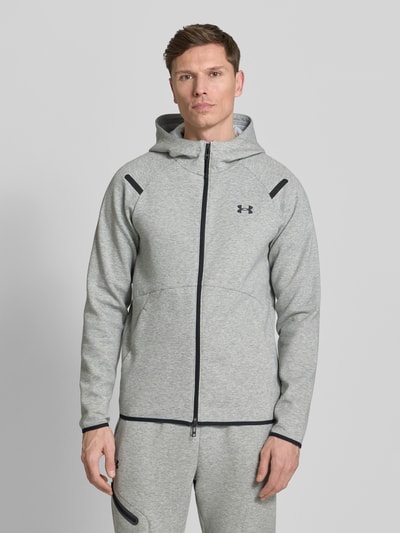 Männermodel trägt Regular Fit Jacke mit Kapuze Modell 'Unstoppable' von Under Armour in Hellgrau mit Logo-Print und Zweiwege-Reißverschluss, Ganzkörperansicht von vorne. Elastischer Material-Mix mit Baumwoll-Anteil, geteilte Känguru-Tasche, elastische Abschlüsse.