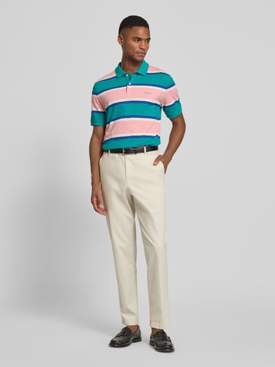 Männermodel trägt Slim Fit Poloshirt mit Logo-Stitching von Gant in Türkis, Pink, Weiß gestreift aus Bio-Baumwolle, Ganzkörperansicht von vorne.