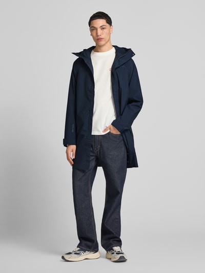 Męski model nosi Regular Fit Regenmantel mit Logo-Detail Modell 'Brussel' od Helly Hansen, kolor granatowy, wpuszczane kieszenie, kaptur ze stójką, długość tyłu w rozmiarze M: 90 cm, szerokość ramion w rozmiarze M: 42 cm, szerokość klatki piersiowej w rozmiarze M: 51 cm, długość rękawów w rozmiarze M: 68 cm, gładka tekstura, pełny widok z przodu