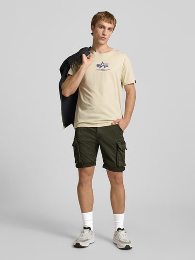 Męski model nosi Shorts mit Cargotaschen Modell 'CREW SHORT' od Alpha Industries, bawełna z elastanem, krój regular fit, kolor khaki, szerokość w rozmiarze 32: 44 cm, długość wewnętrzna nogawki 22 cm, widok całej sylwetki z przodu.