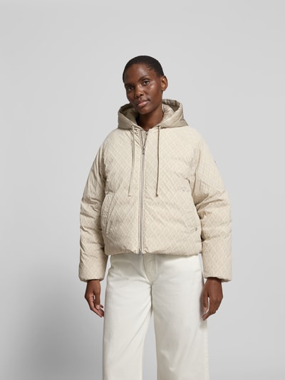 Frauenmodel trägt Regular Fit Jacke mit Allover-Logo-Print von Moose Knuckles, Beige, Elastischer-Material-Mix, Kapuze mit Tunnelzug, Zweiwege-Reißverschluss, Ganzkörperansicht von vorne. Rückenlänge 60 cm, Brustweite 59 cm, Ärmellänge 63 cm, Größe S.