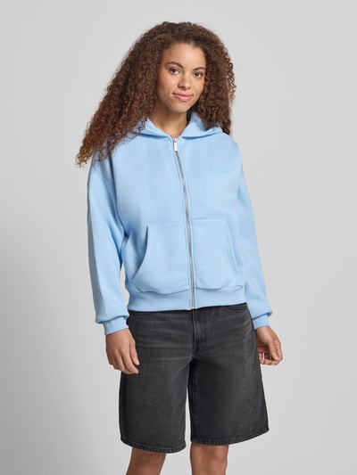 Modelka nosi Relaxed Fit Sweatjacke mit Kapuze od REVIEW z czystej bawełny, z kapturem, suwakiem, nadrukiem logo, kieszeniami kangurkami, krój relaxed fit, jasnoniebieski kolor, miękka faktura, widok całej sylwetki z przodu.