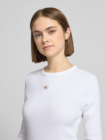 Vrouwenmodel draagt Longsleeve mit Label-Detail van Calvin Klein Jeans in wit, slim fit, geribde ronde hals, katoen-elastaan mix, detailopname bovenlichaam.