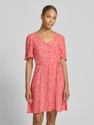 Frauenmodel trägt Minikleid aus Viskose mit kurzer Knopfleiste Modell 'GRACE' von Vila, Taillierter Schnitt, V-Ausschnitt, Bindegürtel, rot mit weißem Muster, Ganzkörperansicht von vorne.