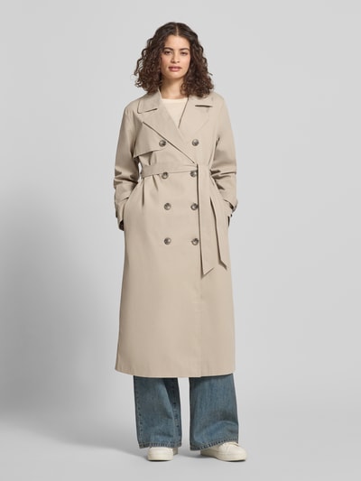 Modelka kobieca nosi Regular Fit Trenchcoat mit Reverskragen Modell 'DISA LIFE' od Only, beżowa, długi rękaw, pasek w talii, krój regular fit, dwurzędowe guziki, boczne kieszenie, gładka faktura, widok całej sylwetki od przodu.