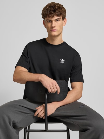 Männermodel trägt T-Shirt mit Label-Stitching von adidas Originals in Schwarz, reine Baumwolle, Regular Fit, Nahaufnahme Oberkörper.