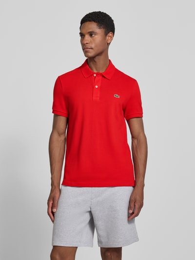 Männermodel trägt rotes Slim Fit Poloshirt aus reiner Baumwolle von Lacoste, weiße Knöpfe, fein strukturierte Oberfläche, Ganzkörperansicht von vorne.