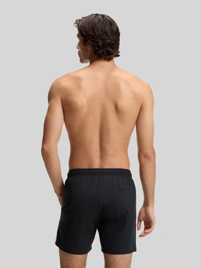 Männermodel trägt Badeshorts mit Tunnelzug Modell 'ICONIC' von BOSS in Schwarz mit Kontraststreifen, Ganzkörperansicht Rückansicht.