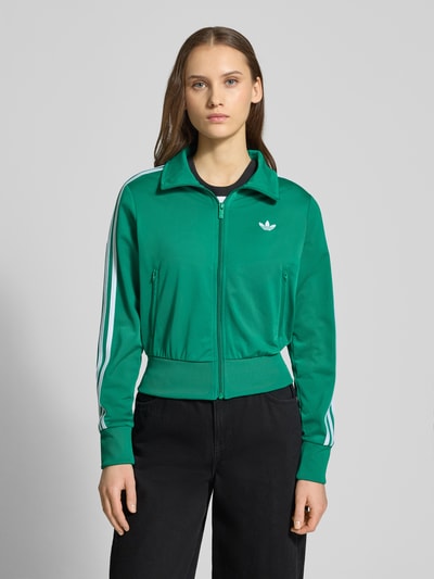 Modelka nosi zieloną Trainingsjacke mit Label-Stitching von adidas Originals, krój regular fit, stójka, kieszenie na zamek, widok na cały przód.