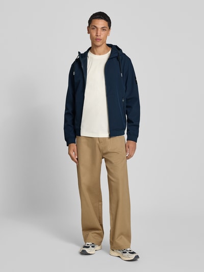 Männermodel trägt Regular Fit Jacke mit Logo-Detail von JACK & JONES in Dunkelblau, helle Hose, neutrale Farben, Ganzkörperansicht von vorne. Herren Jacke, Softshell, Kapuze mit Tunnelzug, Eingrifftaschen mit Reißverschluss, elastische Abschlüsse, Rückenlänge 66 cm, Schulterbreite 46 cm, Brustweite 53 cm, Ärmellänge 67 cm.