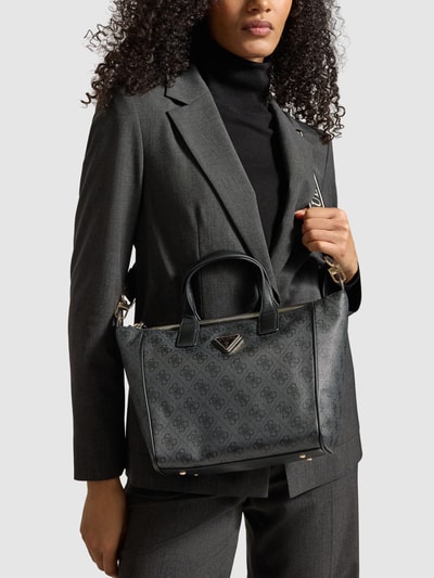 Frauenmodel trägt Tote Bag mit Label-Detail Modell 'FOLLIE' von Guess in Anthrazit mit Logo-Struktur, Tragehenkel und abnehmbarem Schulterriemen, Hauptfach mit Reißverschluss, Innenfach mit Reißverschluss, Maße ca. 35x23x11 cm, Länge Schulterriemen 104 cm, Nahaufnahme Oberkörper.