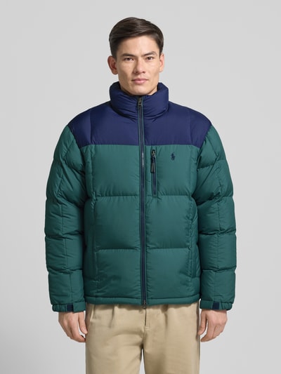 Męski model nosi zielono-granatową Daunenjacke mit Zweiwege-Reißverschluss und Stehkragen von Polo Ralph Lauren, regular fit, detal z logo, widok całej sylwetki z przodu.
