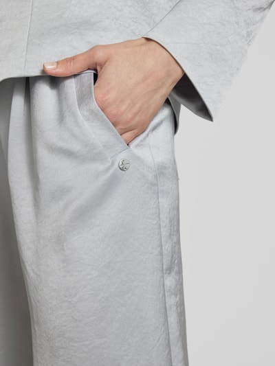 Kobiecy model nosi Wide Fit Stoffhose mit gelegten Falten von Marc Cain, mieszanka acetatu, wpuszczane kieszenie, zamek błyskawiczny, jasnoszary materiał, detaliczny widok boczny, detal wpuszczanej kieszeni, długość wewnętrzna nogawki 83 cm, szerokość w rozmiarze 36: 38 cm, detaliczny widok okolicy kieszeni.
