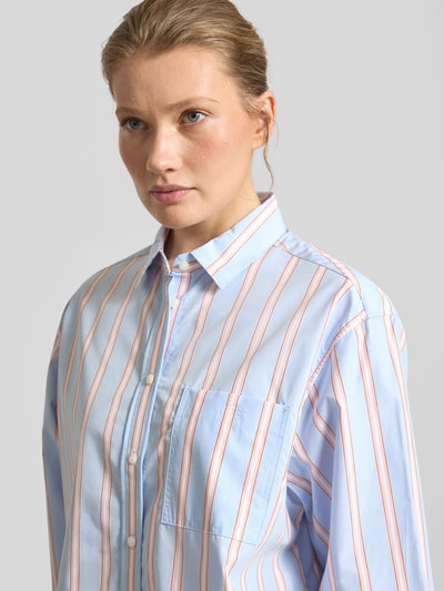 Frauenmodel trägt Oversized Hemdbluse aus reiner Baumwolle von Lacoste, Optik:Allover-Muster, Passform:Oversized, Hemdkragen, Label-Stitching-Applikation, Knopfleiste, Ärmelform:Langarm, Manschette, Brusttasche, Materialverarbeitung:Popeline, Material:reine Baumwolle, gestreifte Struktur in Hellblau und Rosa, Nahaufnahme Oberkörper.