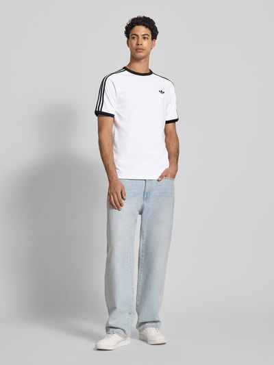 Männermodel trägt T-Shirt mit Label-Stitching von adidas Originals in Weiß und Schwarz, Baumwoll-Mix, Regular-Fit, Ganzkörperansicht von vorne.