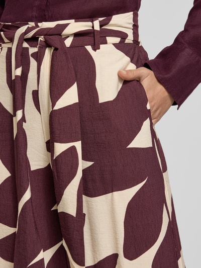 Frauenmodel trägt Stoffhose aus Viskose-Mix mit Bindegürtel Modell 'PLUMAS' von Mango, Allover-Print in Lila-Creme, Nahaufnahme Oberkörper.