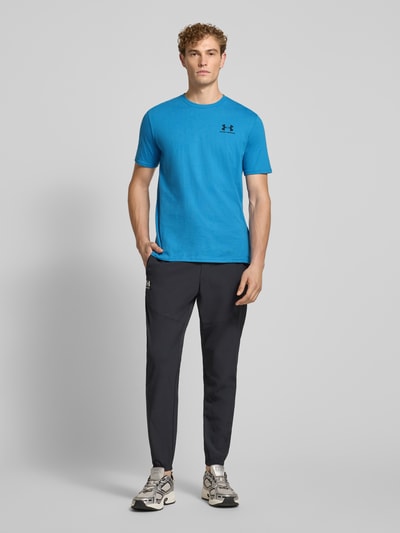 Männermodel trägt Regular Fit Sport-T-Shirt aus Baumwoll-Mix von Under Armour in Blau und dunkler Hose, Ganzkörperansicht von vorne.